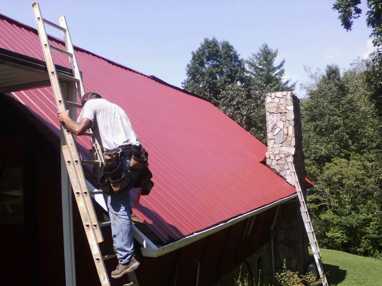 Roofer on a ladder beside a red metal roof and stone chimney — Soffit & Fascia Repair by Roofing Brothers in Nuremberg